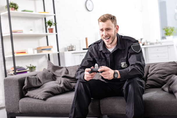 laughing police officer with gamepad sitting on couch and playing video game - Foto, Imagen