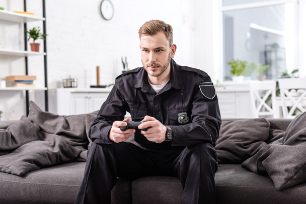 Konzentrierter Polizist mit Gamepad sitzt auf Couch und spielt Videospiel - Foto, Bild
