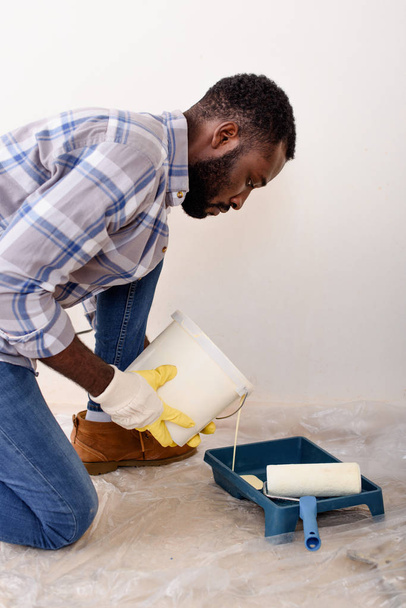 focused african american man in protective gloves pouring paint into roller tray during renovation of home - Foto, Imagem