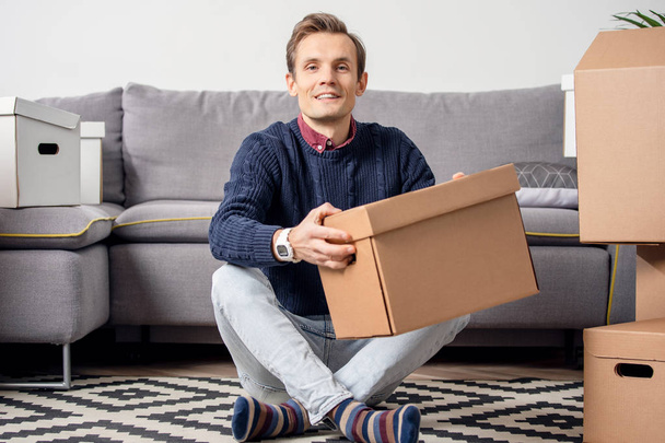 Photo of man sitting on floor with cardboard boxes on background of gray sofa - 写真・画像