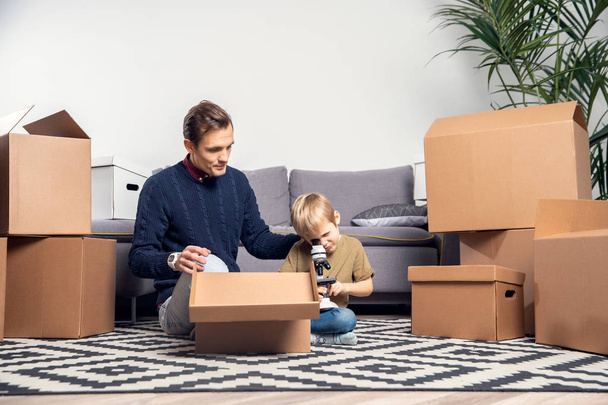 Portrait of man and boy with microscope sitting on floor among cardboard boxes - Foto, immagini