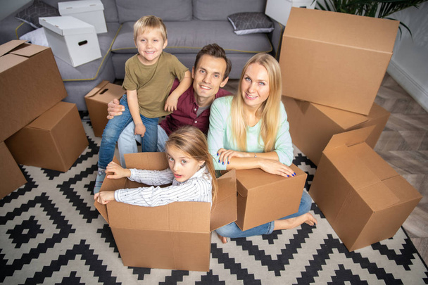 Photo from above young parents and children among cardboard boxes in new apartment - Foto, imagen