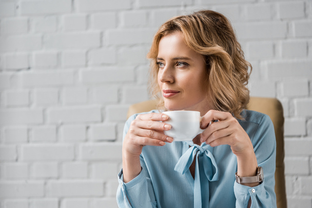 smiling businesswoman holding cup of coffee and looking away in office - Fotografie, Obrázek
