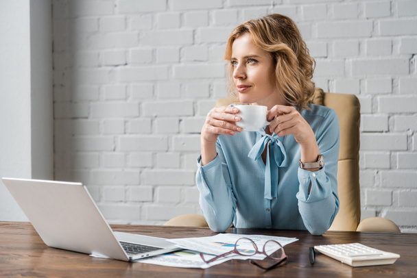 smiling businesswoman holding cup of coffee and looking away while sitting at workplace - Фото, зображення