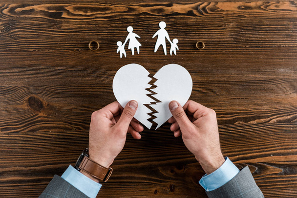 cropped shot of man holding teared heart with family and engagement rings on wooden table, divorce concept - Photo, Image