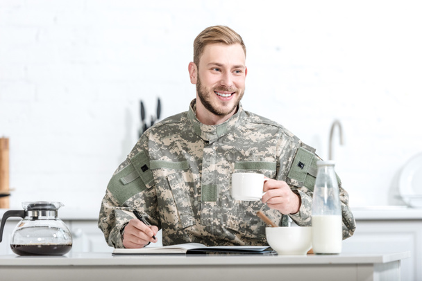 Smiling man in camouflage uniform writing in notebook and having breakfast - Foto, Bild