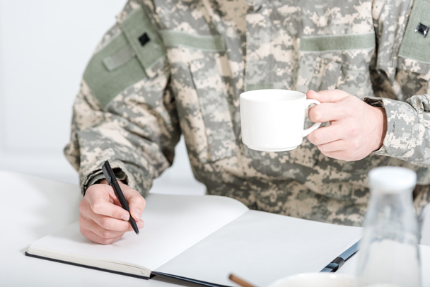 abgeschnittene Ansicht eines Mannes in Militäruniform, der Kaffee trinkt und in Notizbuch schreibt - Foto, Bild