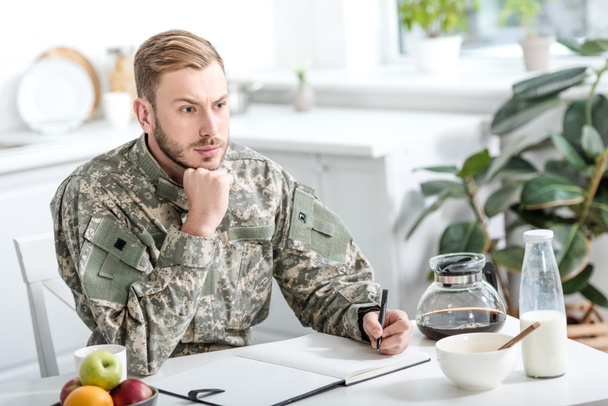 soldado del ejército pensativo sentado en la mesa de la cocina y la escritura mientras desayuna
 - Foto, Imagen