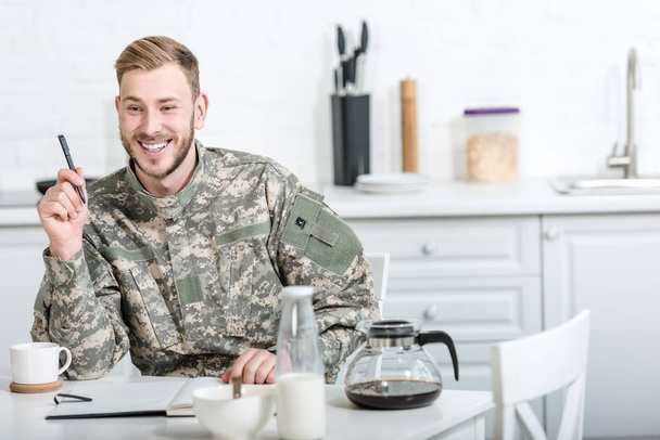 smiling army soldier sitting at kitchen table with pen and notebook while having breakfast - Photo, Image