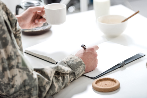обрезанный вид военнослужащего, сидящего за кухонным столом, пишущего и завтракающего
 - Фото, изображение