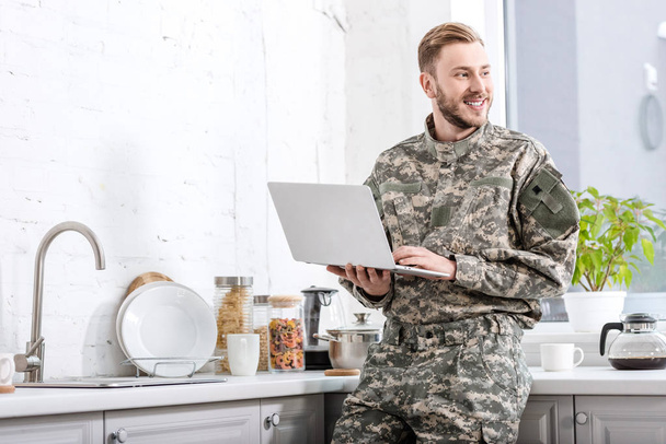 smiling army soldier using laptop at kitchen and looking away - Foto, immagini