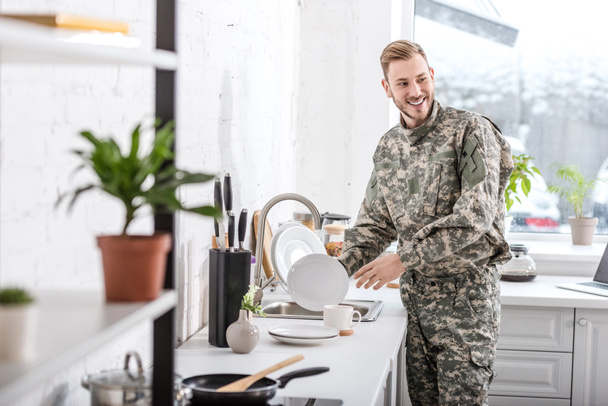 lachende leger soldaat schoonmaken gerechten in de keuken - Foto, afbeelding