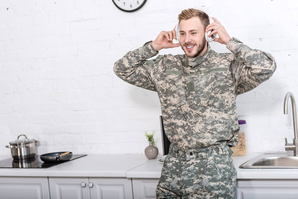 szép katona katonai egyenruhában viselésével fülhallgatók és zene a konyha - Fotó, kép