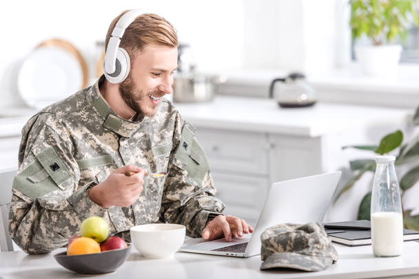 handsome soldier in headphones using laptop at kitchen table while having breakfast - Foto, afbeelding