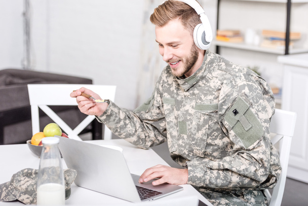 台所のテーブルで朝食をとりながらノート パソコンを使用してヘッドフォンで笑顔の兵士 - 写真・画像