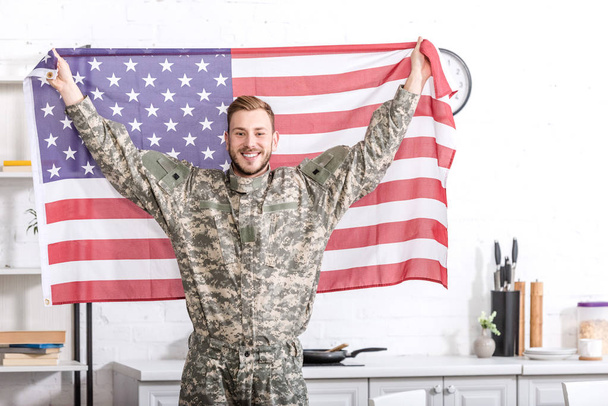 bonito soldado do exército em pé, olhando para a câmera e orgulhosamente segurando bandeira americana
 - Foto, Imagem