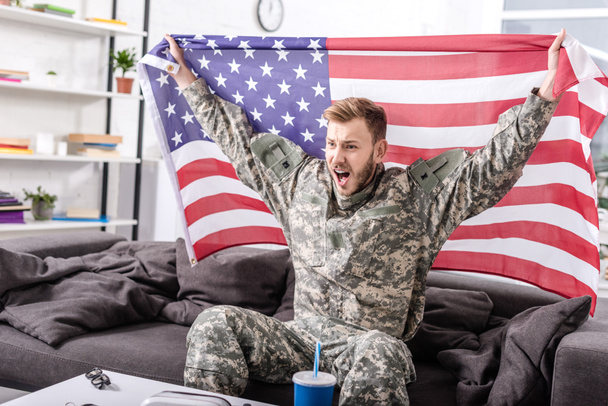 excited army soldier sitting on couch, cheering and proudly holding american flag - Φωτογραφία, εικόνα