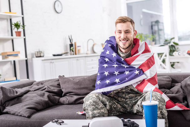 smiling army soldier covered in american flag sitting on couch and watching football match - Photo, Image