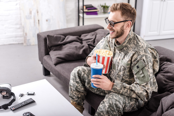 smiling army soldier in 3d glasses on couch watching movie with popcorn and soda water - Photo, Image