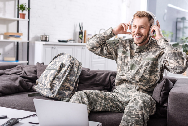 soldat souriant de l'armée dans les écouteurs assis sur le canapé et en utilisant un ordinateur portable à la maison
 - Photo, image