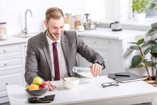 smiling businessman pouring milk in cereal bowl at kitchen table while having breakfast - Φωτογραφία, εικόνα
