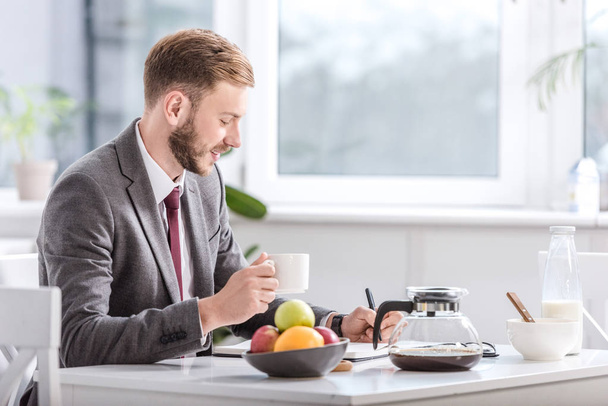 handsome businessman drinking coffee in kitchen and writing in notepad - Foto, imagen
