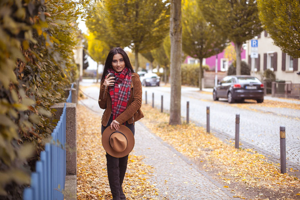Nuori kaunis turkkilainen nainen kävelee eurooppalaisessa kaupungissa. Tytöllä on lasi kahvia. Hyvää ruskeaverikköä hatussa. Syksyllä. Muoti
. - Valokuva, kuva