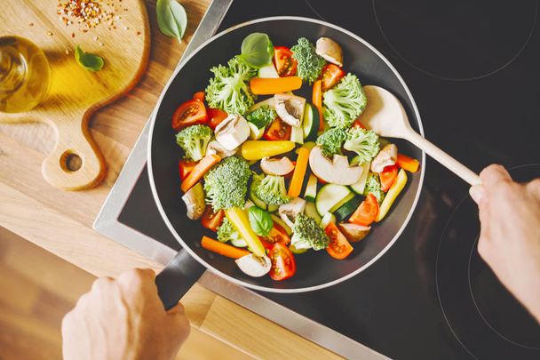 верхний вид человека жарки свежие здоровые овощи с кастрюлей на индукционной плите
 - Фото, изображение