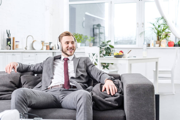 smiling businessman in formal wear wearing headphones and sitting on couch at home - Фото, изображение