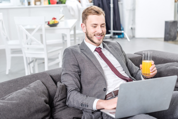 uomo d'affari sorridente in abbigliamento formale bere succo d'arancia e utilizzando il computer portatile
 - Foto, immagini