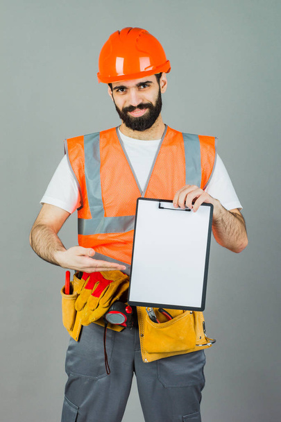 Ein Bauarbeiter mit orangefarbenem Helm auf grauem Hintergrund signalisiert etwas. - Foto, Bild