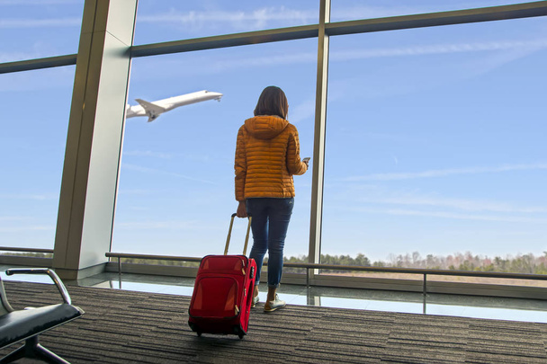Een jonge mooie reiziger vrouw zien het vliegtuig bij het raam van de luchthaven, meisje toeristische met smartphone houdt rode tas en wachten in de hal vliegtuig vertrek. Reizen, vakanties en toerisme concept - Foto, afbeelding