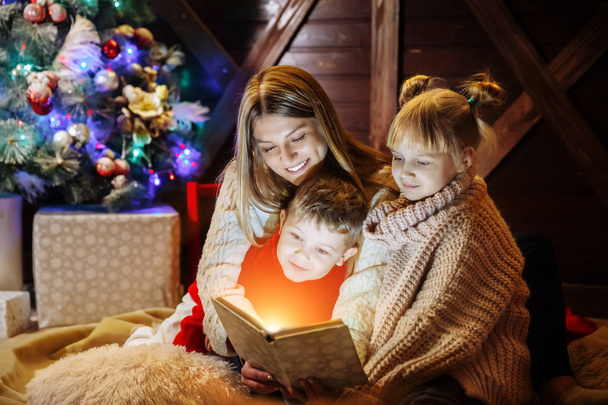 Merry Christmas and Happy New Year. Beautiful family in Christmas interior. Pretty young mother reading a book to her daughter and son near Christmas tree - Foto, immagini