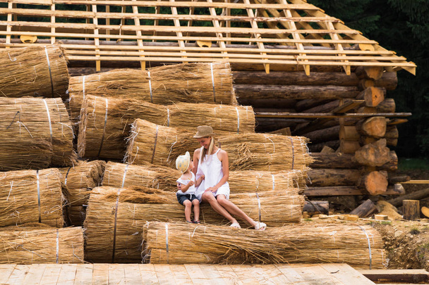 Happy mother with her son sitting on the straw near the wooden house outdoor - Foto, afbeelding