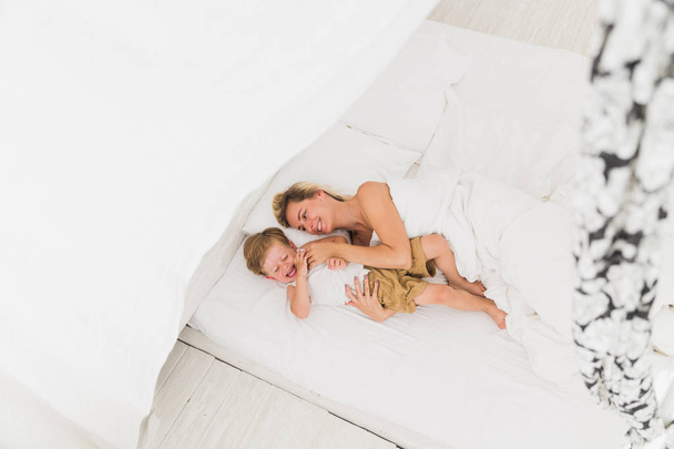 Happy young mother lying on a bed near her little son indoor and they smiling - Foto, Bild