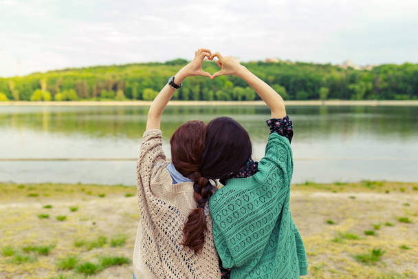 People, friends, sisters and friendship concept - back view, girls, braided hair together, showing heart sign over beautiful landscape on the horizon - Fotografie, Obrázek