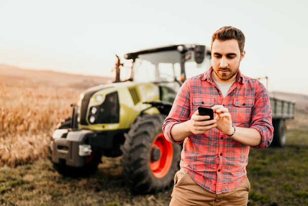 Farmer working and harvesting using smartphone in modern agriculture with tractor background - Фото, изображение