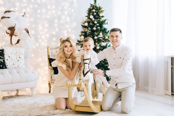 Portrait of a beautiful young family against the backdrop of the Christmas tree. Attractive family celebrates New year, baby riding a white horse and parents smile - Photo, Image