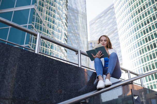 Beautiful woman sitting on the steps and reading a book between skyscrapers - Foto, Imagem