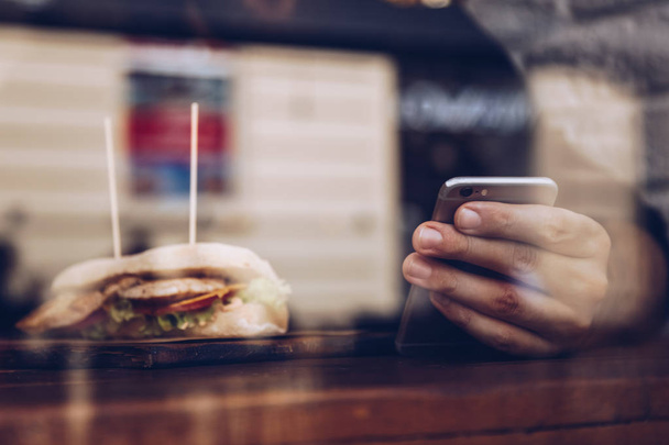 Detailaufnahme, ein Mann in der Mittagspause, der ein gesundes Hühnersandwich isst und in einem Hipster-Restaurant am Fenster tippt. Schuss durch das Glas, in dem sich die Stadt spiegelt. - Foto, Bild