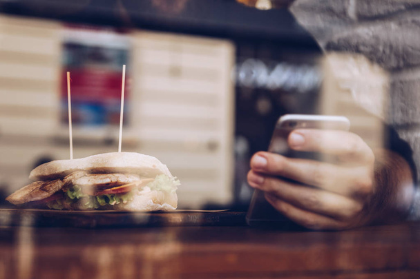 Detailaufnahmen zeigen einen Mann, der in einem kleinen Restaurant ein gesundes Hühnersandwich isst und auf seinem Smartphone tippt. in der Mittagspause, neben dem Fenster sitzend und durch dieses geschossen, mit Stadtreflexionen im Glas. - Foto, Bild