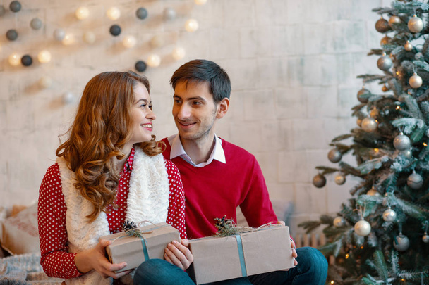 Happy νεαρό ζευγάρι με τα Χριστούγεννα παρουσιάζει ψάχνει με το άλλο σε ένα στολισμένο δέντρο φόντο - Φωτογραφία, εικόνα