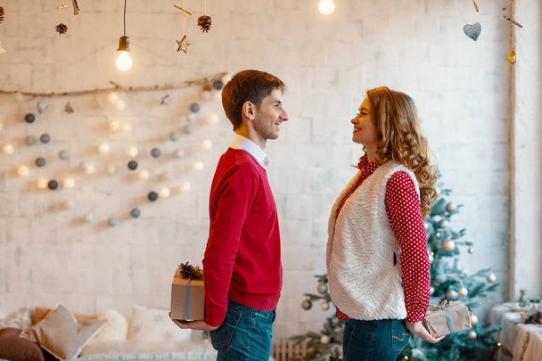 Чоловік і жінка ховають різдвяні подарункові коробки позаду, дивлячись один на одного з різдвяними прикрасами на фоні
 - Фото, зображення