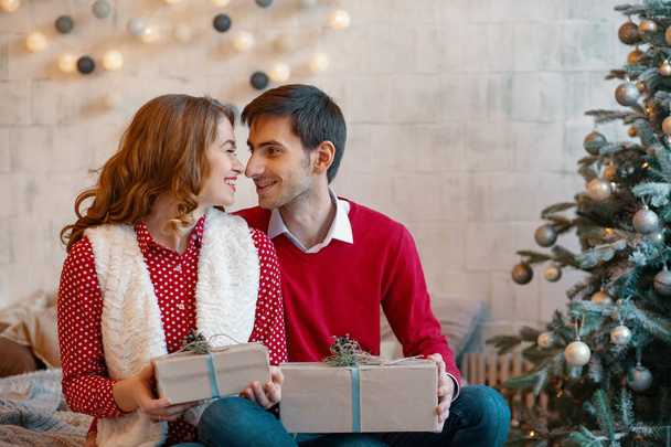Счастливая молодая пара с рождественскими подарками смотрят друг на друга на декоративном фоне елки
 - Фото, изображение