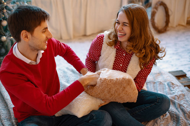 Heureux couple caucasien aimant devient fou le soir de Noël près de l'arbre du Nouvel An, ils sont assis sur le lit, riant beaucoup, se battant avec des oreillers
 - Photo, image