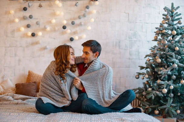Young man looking to in to his girlfriend eyes smiling sitting with crossed legs on a bed with decorated tree and window on background. Happy loving couple enjoying Christmas. - Photo, image