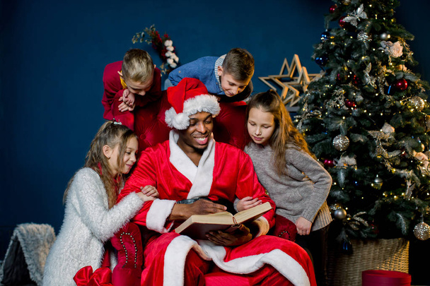 Santa Claus reads an ancient book with Christmas stories for children sitting around a red chair - 写真・画像
