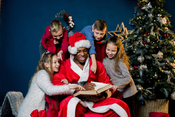 Santa Claus reads an ancient book with Christmas stories for children sitting around a red chair - Foto, Imagen