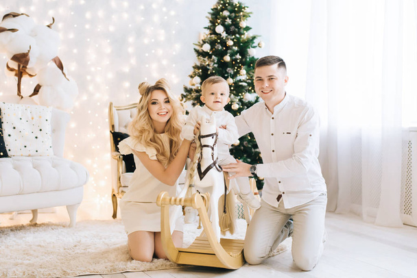 Portrait of a beautiful young family against the backdrop of the Christmas tree. Attractive family celebrates New year, baby riding a white horse and parents smile - Zdjęcie, obraz