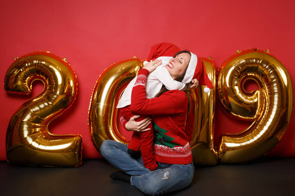 Buon Babbo Natale mamma ragazzo in cappello di Natale che celebra festa isolata su sfondo brillante parete rossa dorata brillantini numeri palloncini d'aria full length studio ritratto. Felice anno nuovo concetto
 - Foto, immagini